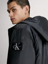 Campera cortavientos negra con capucha, confeccionada en poliéster reciclado. Cuenta con cierre de cremallera, puños y bajo elásticos, bolsillos laterales y logo de Calvin Klein en la manga.