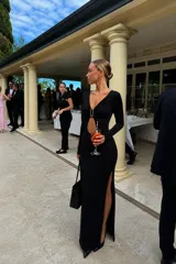 Vestido largo negro de manga larga con escote profundo y detalle de hebilla metálica en el centro del pecho. Presenta una abertura frontal tipo cut-out y una abertura lateral en la falda.