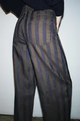 Jeans de tiro medio con corte barrel leg, confeccionados en denim con rayas verticales a contraste en tonos azul oscuro y marrón/tostado. Presentan cinco bolsillos y cierre frontal con cremallera y botón.