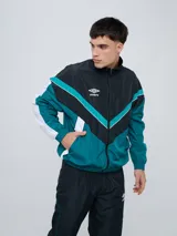 Campera deportiva Umbro modelo Beat, confeccionada en nylon ligero con diseño de bloques de color en blanco, azul marino y verde azulado. Presenta cierre frontal, cuello alto y elásticos en puños y cintura.