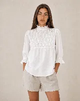 Blusa blanca holgada de lino con detalle de nido de abeja hecho a mano en el delantero, cuello alto con volados, mangas tres cuartos con puños elásticos fruncidos y cierre trasero con botones forrados.