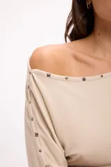 Remera de manga larga color beige con diseño de hombro caído y detalle de tachas metálicas a lo largo del escote.