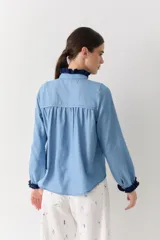 Camisa de jean azul claro con detalles de crochet en color azul oscuro en el cuello, la botonera frontal y los puños. Presenta un corte recto, mangas largas y botones de madera.