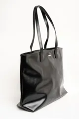 Bolso tipo tote bag color marrón, confeccionado en cuero sintético, con doble asa superior y detalle de placa metálica dorada con logo en la parte frontal.