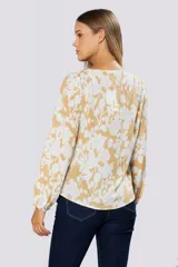Blusa de manga larga con estampado floral en tonos blanco y mostaza. Presenta escote en V con botones frontales, frunces en los hombros y puños abotonados con detalle de pin metálico con logo.