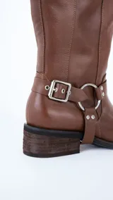 Bota de caña alta confeccionada en cuero color suela, con diseño de estilo western. Presenta tres hebillas metálicas en la parte superior de la caña y un estribo con argolla metálica en el tobillo. Cuenta con taco cuadrado de altura media.