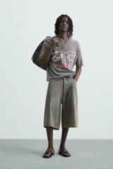 Camiseta gris de punto con efecto lavado y roturas, corte "boxy fit", cuello redondo y manga corta. Presenta un estampado frontal en blanco y rojo con diseño abstracto.