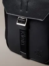Morral Calvin Klein modelo Cargo Reporter, color negro, confeccionado en poliuretano con diseño estructurado, solapa frontal y cierre de hebilla. Cuenta con correa de hombro ajustable y detalles de la marca grabados.