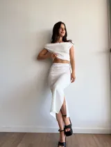 Conjunto de dos piezas en color blanco, compuesto por un top con escote bote y hombros descubiertos, y una falda midi con tajo lateral. Ambas prendas están confeccionadas en tela bambula elastizada con textura arrugada.