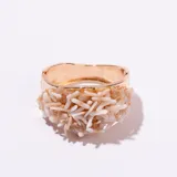 Brazalete tipo esclava de metal dorado, con un diseño ancho y textura acanalada en la parte interna. La parte externa está decorada con múltiples piezas irregulares de color blanco hueso o coral claro, sujetas con alambre fino dorado, simulando ramas o corales.