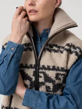 Chaleco de polar con diseño jacquard de motivos geométricos en tonos beige y gris oscuro. Presenta cuello alto, cierre frontal metálico con bordes de cuero sintético y bolsillos laterales con cierre.