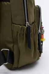 Mochila de nylon negra con asas superiores reforzadas y tiras regulables. Cuenta con un bolsillo frontal con cierre metálico y un detalle colgante de cuentas de colores. Los laterales tienen bolsillos elastizados y el compartimento principal tiene cierre.