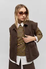 Chaleco de jean color marrón de corte relajado, con hombros caídos, cuello camisero, cierre frontal con botones y bolsillos delanteros grandes aplicados.