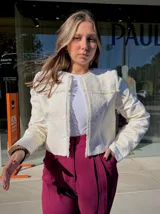 Chaqueta corta de estilo estructurado en color beige, con diseño de brocado floral en relieve y terminaciones desflecadas en el frente y cuello. Presenta mangas largas y cierre frontal oculto.