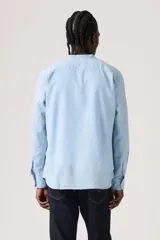 Camisa celeste de manga larga con cuello mao y cierre frontal de botones. Cuenta con un bolsillo aplicado en el pecho adornado con una pequeña etiqueta roja.