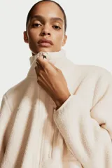Campera deportiva color crema en tejido sherpa, de corte boxy, con cuello elevado, cierre frontal, bolsillo superior con cierre, bolsillos laterales insertados con cierre, elástico fino en los puños y cordón de ajuste en el bajo.
