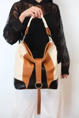 Bolso tipo hobo confeccionado en cuero vacuno, con diseño tricolor en tonos arena, caramelo y negro con textura croco. Posee una manija corta, una correa larga ajustable y desmontable, y detalles de costuras artesanales con flecos decorativos en la parte frontal.