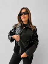 Chaqueta tipo biker de eco cuero negro con diseño estructurado. Presenta apliques de tachas metálicas en formas orgánicas sobre paneles de gamuza sintética, cierre frontal metálico y bolsillos laterales con cierre.