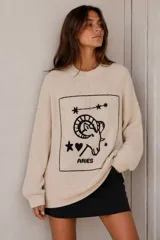 Sweater de punto color crudo, con cuello redondo, mangas largas y diseño oversize. Presenta un parche frontal rectangular con bordado a mano de la constelación y el carnero del signo Aries, acompañado de detalles de estrellas, un corazón y la palabra Aries.