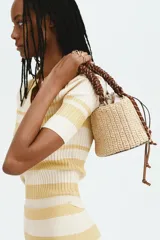 Cartera tipo tote de paja trenzada, con estructura rígida y base redonda. Cuenta con una bolsa interior de lona color crudo con cierre ajustable mediante cordones de cuero. Posee una manija superior de cuerda marrón trenzada y detalles metálicos dorados.