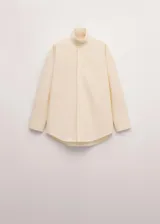 Blusa oversize de algodón en color marfil, con cuello camisero alto, manga larga con puños abotonados y cierre frontal de botonadura oculta.