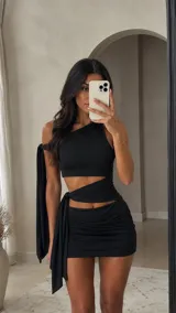 Vestido corto negro con diseño asimétrico, escote de un solo hombro y aberturas laterales en la cintura con detalle de lazo lateral.