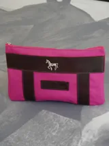 Sobre tipo cartuchera de lona naranja con detalles y apliques en cuero marrón. Presenta un aplique metálico en forma de caballo y un parche con el logo de la marca grabado en el frente.