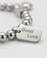 Pulsera elástica de acero compuesta por pequeñas esferas metálicas. Presenta dos dijes colgantes: uno en forma de corazón y otro rectangular con la inscripción 'Good Luck'.