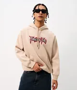 Buzo canguro beige de algodón fleece con capucha y cordón ajustable. Presenta un bordado frontal con el texto "Forged by Fire" en tipografía estilo gótico con bordes rojos y negros.