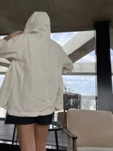 Sobre camisa color crema con capucha, cierre frontal con botones a presión, bolsillos con solapa en el pecho y mangas largas con puños abotonados.