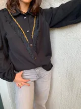 Camisa negra de tejido fino con cuello clásico, cierre frontal con botones, mangas largas abullonadas con puños elásticos y detalle de galón dorado y flecos negros en el frente.