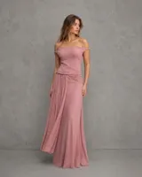 Vestido largo color malva con escote barco y tirantes caídos sobre los hombros. Presenta un cuerpo ajustado y falda fluida con un drapeado fruncido en la cintura que cae hacia un lado.