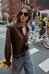 Camisa marrón de manga larga con cuello solapa y cierre frontal de botones. Los puños terminan con lazos anudados.