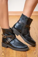 Bota corta de cuero negro con acabado brillante, suela plana de goma y detalle de tiras cruzadas con hebillas metálicas laterales. Cuenta con cierre lateral para facilitar el calce.
