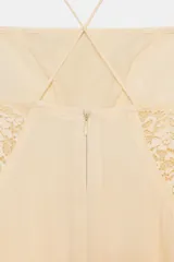 Vestido corto color crema/hueso, con escote recto y tirantes finos cruzados en la espalda. Presenta paneles de encaje en el centro del busto y a los lados de la cintura. La falda es fluida con godets y bajo asimétrico.