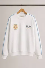 Buzo de cuello redondo en color blanco, con diseño bordado que incluye un sol y estrellas. Presenta líneas verticales en color celeste a lo largo de las mangas y puños acanalados.