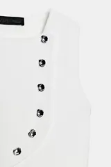 Top de punto blanco sin mangas, de cuello redondo y corte ajustado. Presenta un diseño asimétrico con una solapa cruzada decorada con una hilera de botones metálicos oscuros dispuestos en diagonal que se extienden hasta el dobladillo asimétrico.