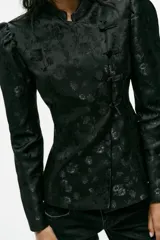 Blazer entallado de color negro con diseño jacquard floreado, cuello mao, manga larga abullonada y cierre frontal cruzado con alamares.