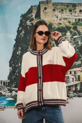 Cardigan oversized de punto grueso, color crema con franjas horizontales rojas. Presenta cuello redondo, puños y dobladillo con ribete a rayas marrones y celestes. Cierre frontal con botones marrones y logo bordado en el pecho.