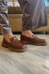 Mocasín de cuero color suela con punta cuadrada y detalle de tachas metálicas en la faja frontal. Cuenta con suela de caucho con vira a la vista.