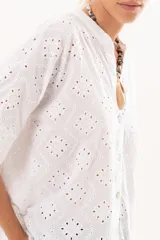 Camisa de broderie blanca con silueta oversize, escote en V, cierre frontal con botones y mangas 3/4. Presenta un diseño con bajo asimétrico, siendo ligeramente más larga en la parte posterior.