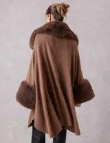 Ruana de tejido color tostado con cuello, bordes frontales y puños amplios con apliques de piel sintética al tono. Presenta una abertura central con cierre de botón tipo trenca.