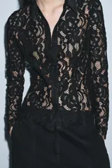 Camisa de manga larga confeccionada en encaje negro con patrón floral. Presenta cuello solapa de tejido liso negro y cierre frontal de botones. El bajo del ruedo y los puños tienen terminación de encaje festoneado.