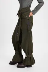 Jean tipo baggy de pierna ancha y recta, color marrón oscuro. Presenta detalles de costuras decorativas, bolsillos laterales redondeados y bolsillos traseros grandes. Incluye tiras ajustables con hebillas metálicas en los tobillos para un efecto fruncido.