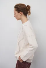Buzo de punto color beige con diseño de trenzas y costuras decorativas en contraste que forman rombos. Presenta cuello redondo, mangas largas y puños acanalados.