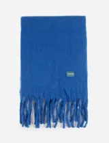 Bufanda de textura suave y acabado afelpado, color negro, con flecos en los extremos y un pequeño parche cuadrado decorativo con logo en color verde y azul.