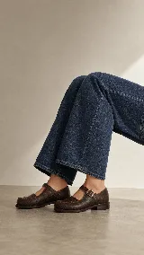 Mocasín de cuero color marrón con acabado texturizado, diseño de punta redonda con costuras artesanales visibles y cierre mediante una tira con hebilla lateral.