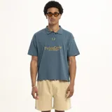 Remera tipo polo de manga corta, color azul verdoso, con cuello y abertura frontal corta. Presenta un bordado en color dorado en el pecho con el texto "PulauCrew" y una frase debajo.