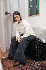 Blusa de color manteca con textura acanalada, cuello redondo y mangas largas con puños acampanados. El diseño presenta múltiples volados horizontales distribuidos a lo largo del cuerpo y las mangas.
