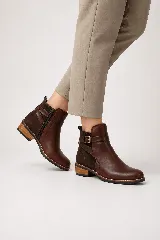 Botines de cuero marrón con diseño de inspiración ecuestre, combinando cuero liso en la parte delantera y gamuza en el talón. Cuentan con una tira decorativa con hebilla metálica alrededor del tobillo, cierre lateral interno, punta redondeada y tacón bajo de madera.